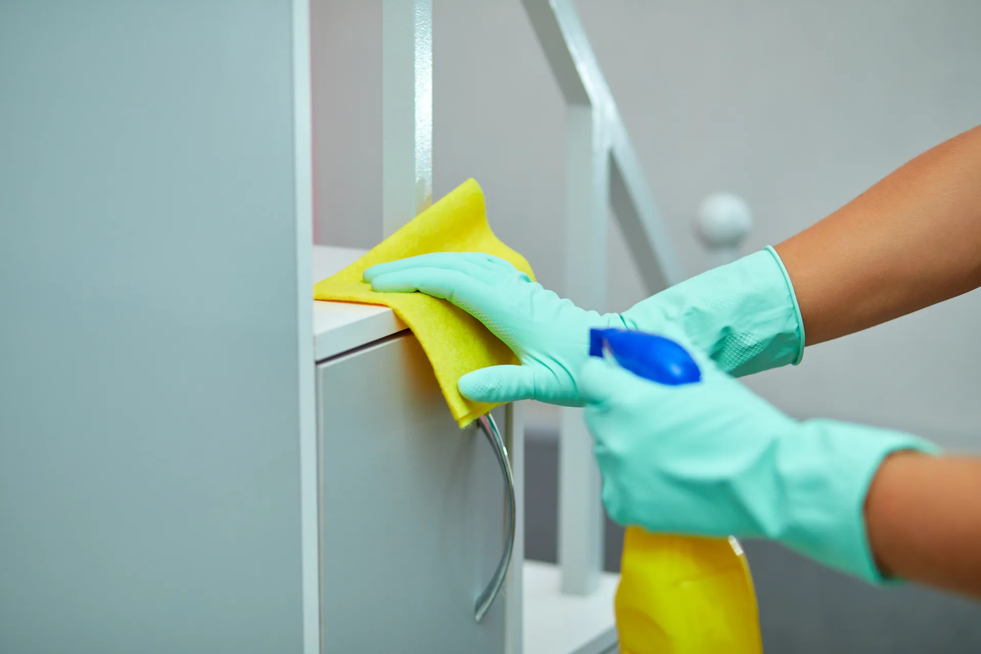Gloved hands wiping a cabinet front with a microfibre cloth and a spray bottle